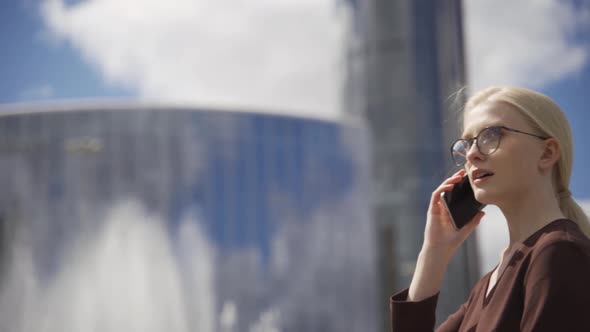 A Business Woman Walks Past Highrise Buildings and Speaks on the Phone alt