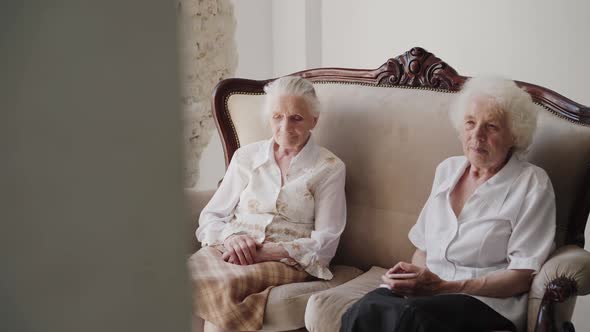 Two Elegant Grandmothers Sitting on a Sofa in Light Room alt