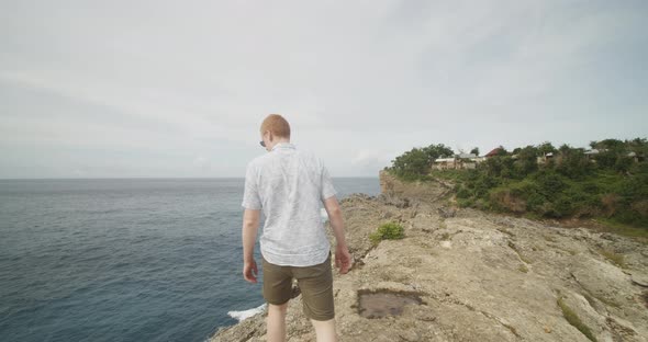 Close Up Following Shot of a Man with Ginger Hair Walking on the Cliff Above the Ocean Alongside alt