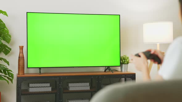 Woman sit on sofa and holding joy game with TV green screen in living room with tree and lamps. alt