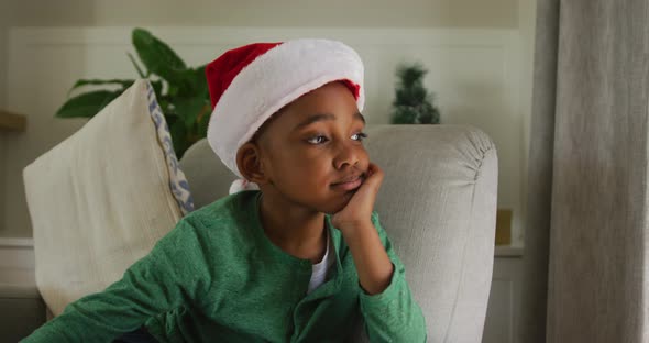Animation of sad african american boy wearing santa hat sitting on sofa at christmas time alt