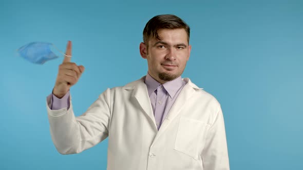 Pleased Man in Medical Coat Twists Removed Protective Mask on Finger and Throws It on Floor alt