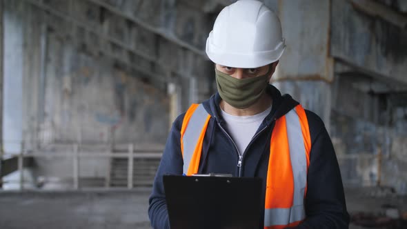 Engineer in the Construction Helmet and Antiviral Protective Mask Inspects a Building or Object