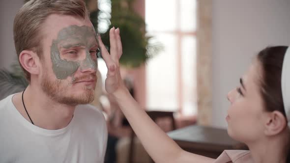 Happy Couple Young Woman Applying Facial Skin Care Mask on Man Boyfriend Face. Cosmetic Beauty