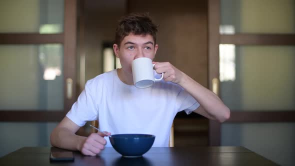 A teenager eats porridge and drinks tea, looking out the window alt