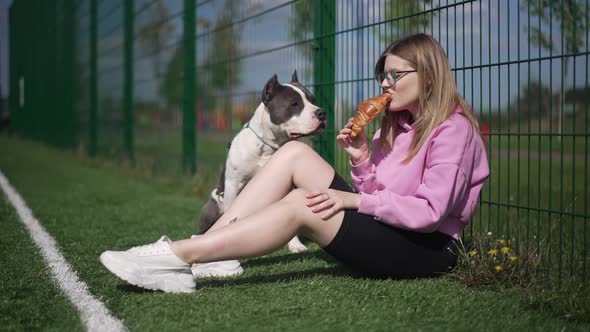 Side View Satisfied Sportswoman Eating Delicious Croissant Sitting at Mesh Fence As American alt