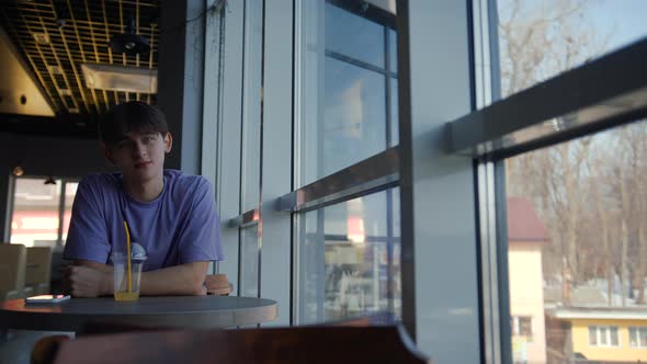 A Young Guy is Sitting in a Cafe By the Big Window Drinking Juice and Smiling