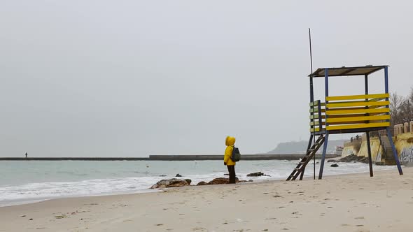 A Man in a Warm Yellow Jacket with a Backpack Stands on the Seashore and Looks Into the Distance