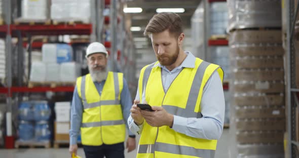 Aged Warehouse Manager Giving Helmet to Young Worker Standing in Using Smartphone alt
