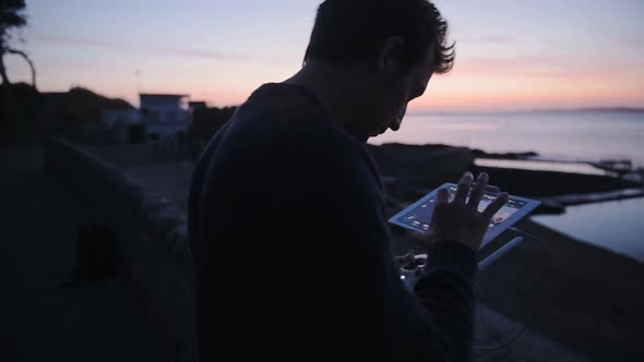 Man piloting a drone with a controller, above outdoor swimming pools, Guernsey seaside, at dusk alt