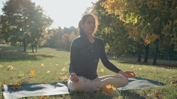 Young Woman Sitting on Mat in Lotus Position in City Park on Sunny Autumn Day alt