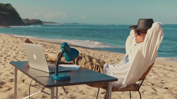 A Young Man Sits on the Beach with Laptop, Resting Putting His Legs on the Table alt