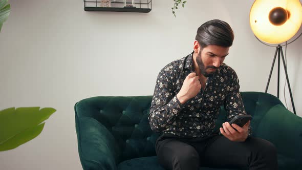 Latin Handsome Bearded Man Sitting on a Greencolored Couch Looking at His Mobile Phone and Cheering alt