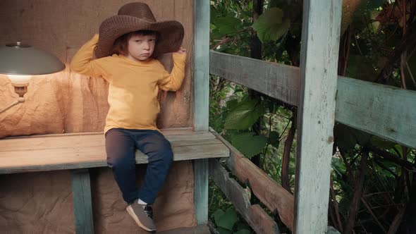 Small Funny Child in Big Cowboy Hat is Sitting on Wooden Porch of Country House