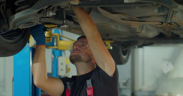 Car Mechanic Is Working Under a Vehicle on a Lift in Service. Repairman Is Using Ratchet. Specialist alt