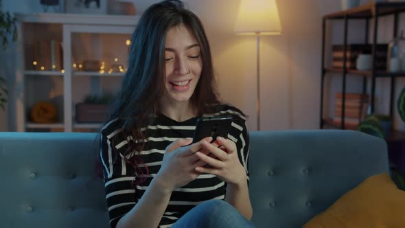 Happy Student Laughing and Using Smartphone Sitting on Couch at Home at Night alt