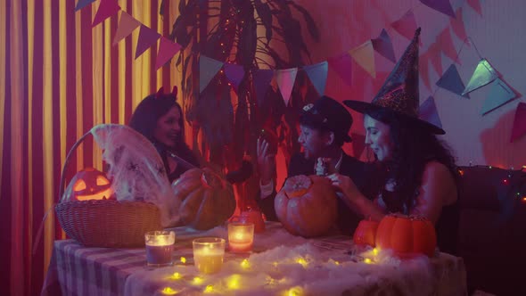 Two Girls and a Guy in Creepy Halloween Costumes are Sitting at a Table Chatting and Carving Faces alt