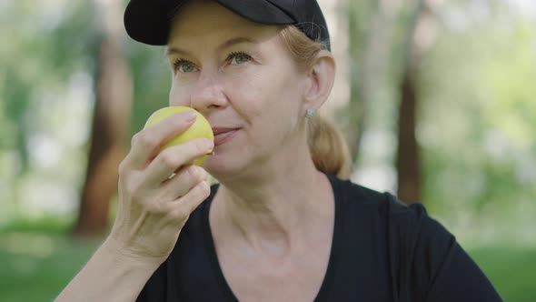 Close-up of Happy Caucasian Woman in Sportswear and Cap Smelling Tasty Apple in Summer Park alt