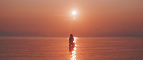 Silhouette of a young woman standing in the sea playfully touching the water with her hands alt