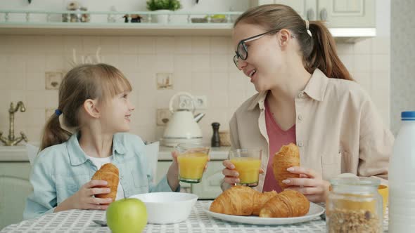 Mother and Daughter Having Breakfast Eating Croissants and Clinking Juice Glasses at Home alt