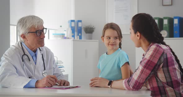 Aged Male Pediatrician Doctor Talking to Little Girl and Mother in Office of Medical Clinic alt