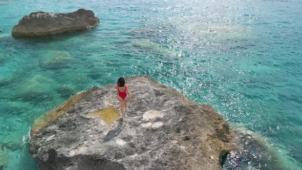 Beautiful Woman in Red Swimsuit Walking Buy Big Rock Looking at the Sea alt