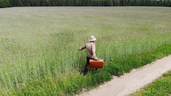 Man Alone Walking By Country Road in a Green Field, Touching Growing Rye, Holding Suitcase in a Hand
