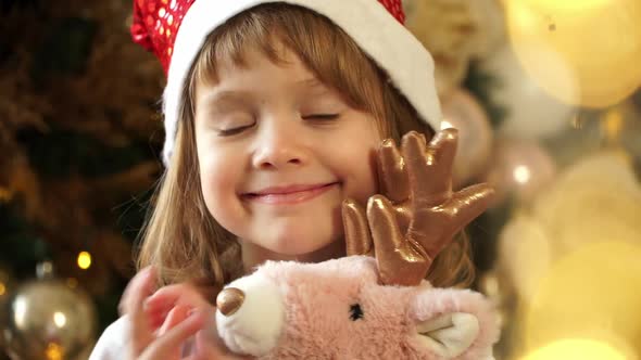 Little Girl in Red Santa Hat Looks at Camera Dances and Smiles on Background Christmas Tree alt