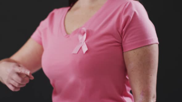 Mid section of a woman pointing to the pink ribbon on her chest against black background alt