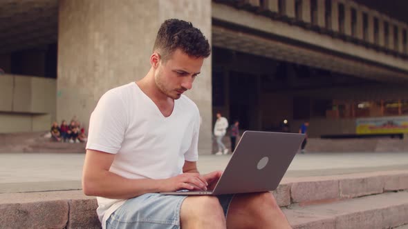 Young Handsome Man in White Tshirt Works with Laptop in the City Center While Sitting on the Steps alt