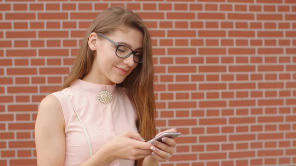 Girl Student in Glasses Smiling and Using Her Phone Against a Brick Wall Background alt