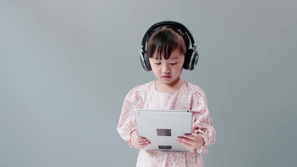 ittle Asian girl with headphone enjoy watching video online on tablet and dancing happily. alt