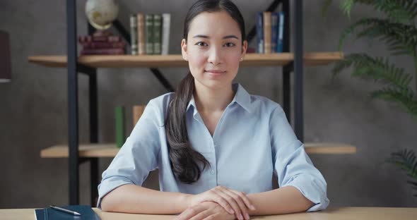 Smiling Young Asian Woman Teacher Sitting at the Table and Looking at Camera alt