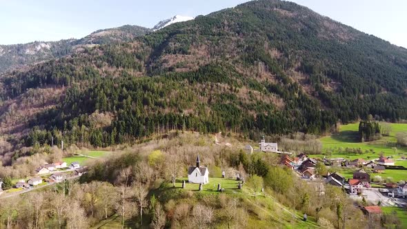 Drone Shot (facing forwards,ing forwards) of a Chapel on top of a Hill in front of Alpine Mountains alt