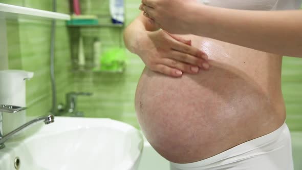Pregnant Girl Lubricates Stomach with Cream Against Stretch Marks in the Bathroom alt