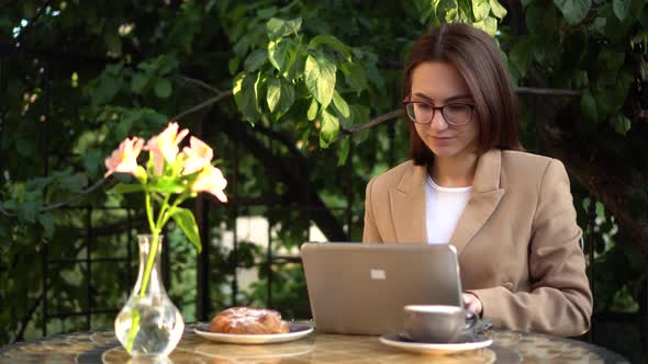 Young Business Woman Sits in a Cafe and Works at a Laptop. Girl with Coffee and a Bun on the Summer alt