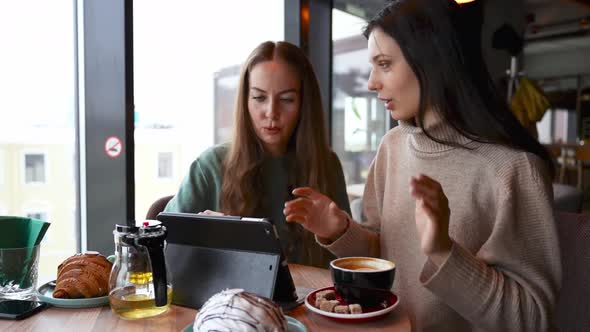 Two Young Beautiful Girls in a Cafe are Looking at a Tablet