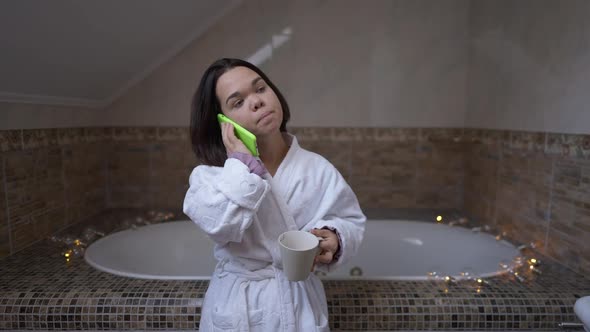 Confident Young Little Woman Talking on Phone Drinking Coffee Standing in Bathroom at Home Indoors alt
