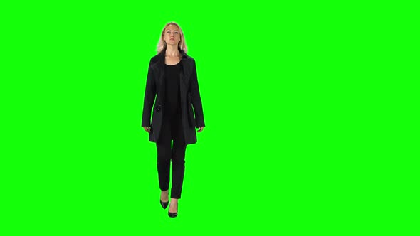 Blonde Girl in Black Coat, Trousers and High-heeled Shoes Going Against a Green Screen. alt