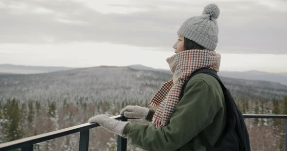 Female Asian Tourist with Backpack Looking at Breathtaking View of Winter Pine Forest and Mountains alt