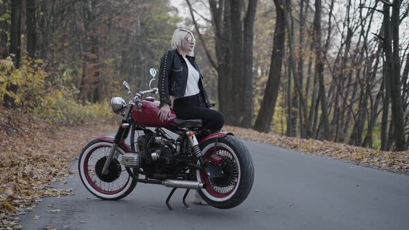 Portrait of Blonde Poses and Corrects Sunglasses at Motorbike on Roadway alt