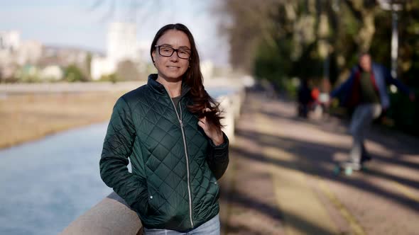 Charming Middleaged Woman in Jacket is Standing on River Embankment in Small Town alt