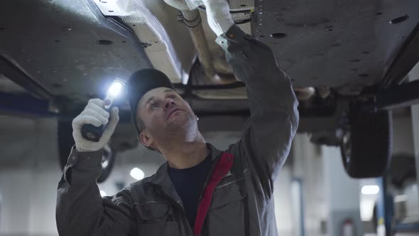 Young Caucasian Man in Workwear with Flashlight Examining Car Bottom. Portrait of Serious alt