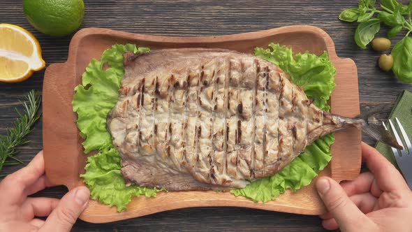 Hands are Putting a Plate with a Grilled White Fish on the Table with Lemons alt