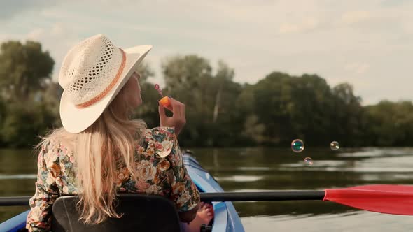 Girl Enjoying At Sunset. Cheerful Blonde Woman Blowing Soap Bubbles. Woman In Hat Relaxing On Kayak alt