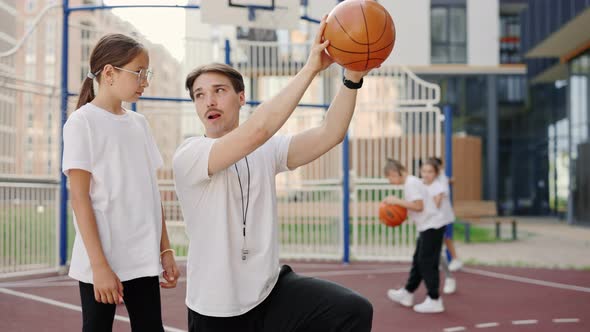Handsome Male Coach Explaining to Child How to Put a Ball Into the Basket on Court Near School alt