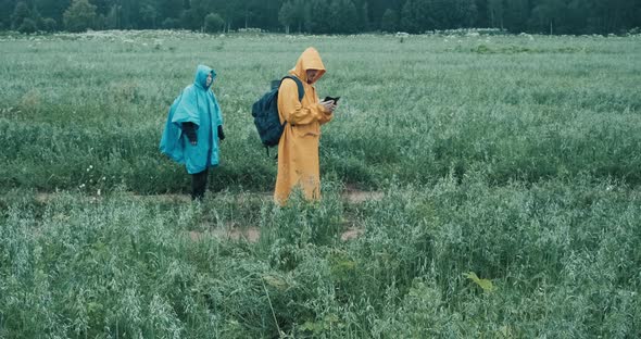 Man and Woman in Raincoats are Walking Through Field and Looking at Map in Phone