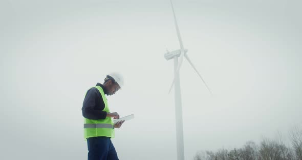 A Man in a Work Uniform is Diagnosing a Wind Turbine Using a Digital Tablet alt