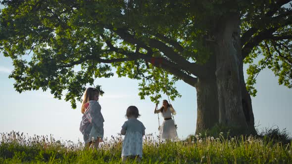 Two Daughters Walking Along Green Field To Their Mother, Swinging on a Rope Swing Tied To Old Tree. alt
