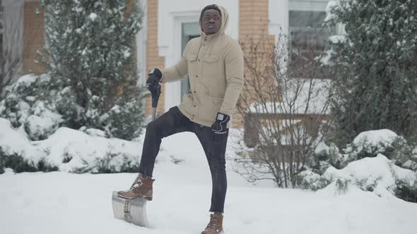Wide Shot Portrait of Thoughtful Young African American Man Standing with Shovel Outdoors on Snowy alt